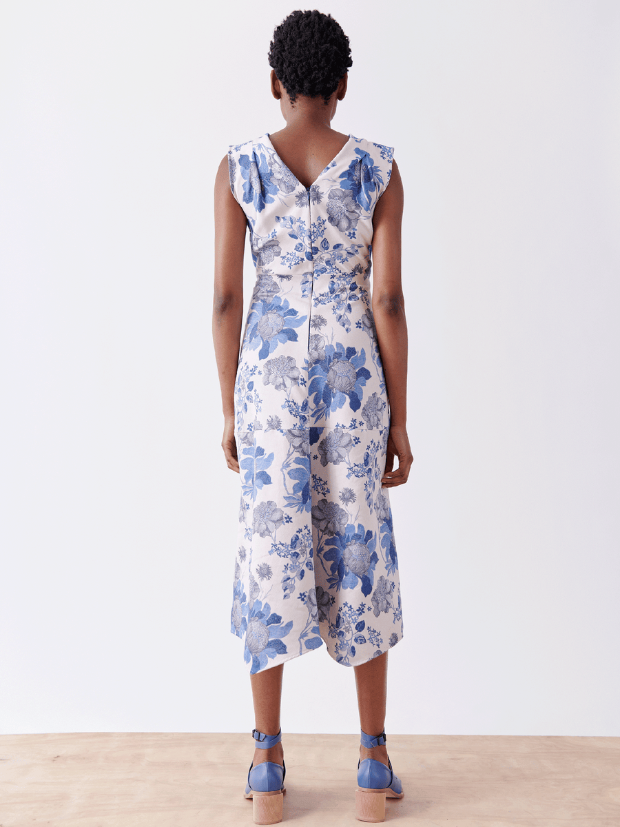 A person showcases the Joi Dress - Final Sale by Zero + Maria Cornejo, featuring a sleeveless floral jacquard design in blue with a V-shaped back and below-knee length. Paired with matching blue sandals on a white background, the look combines elegance and simplicity effortlessly.