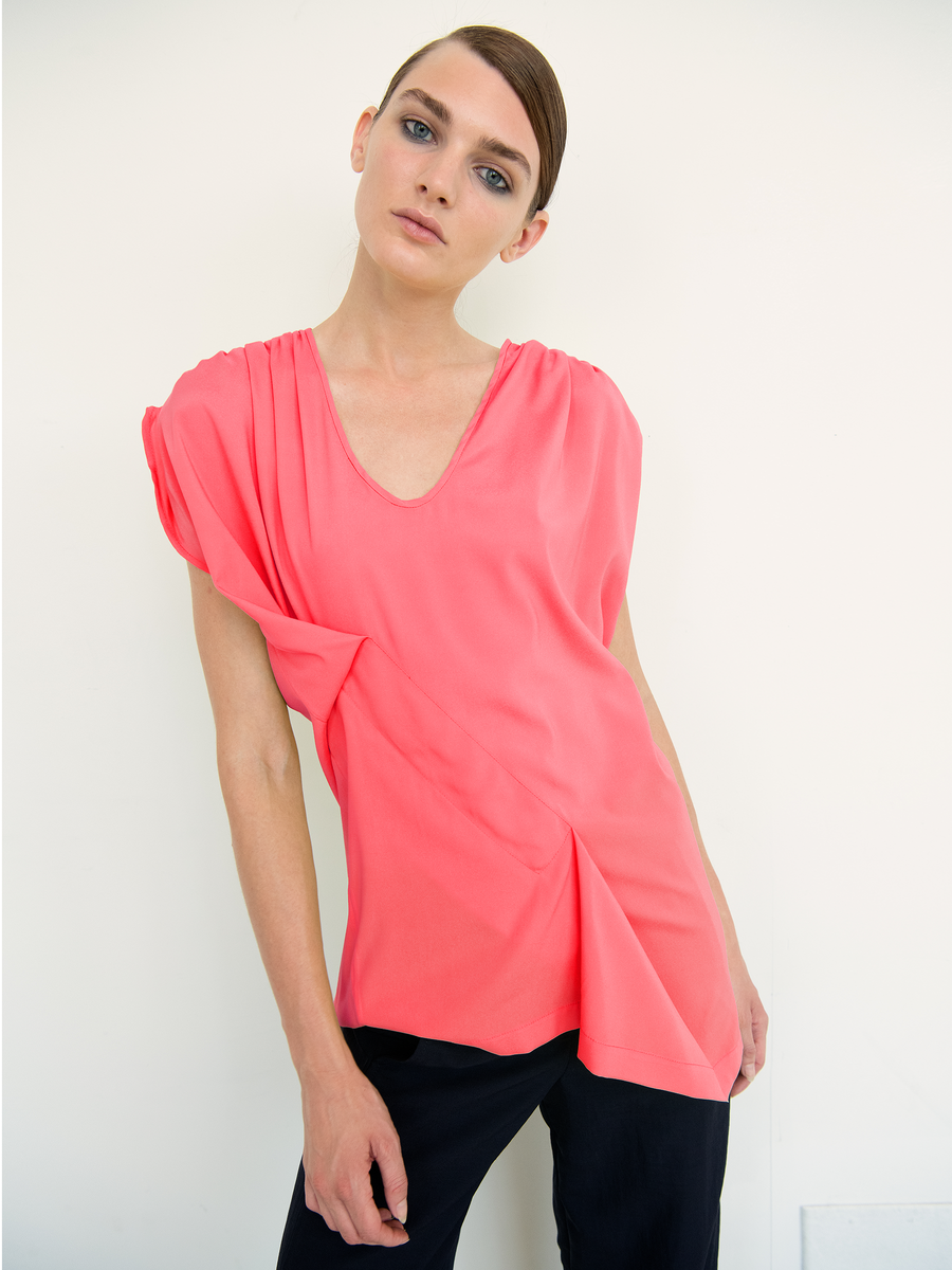 A person models the Ruched Fold Top - Final Sale by Zero + Maria Cornejo, featuring coral pink, short sleeves and a loose, asymmetrical folded front with a V-neck. Paired with black pants against a white backdrop, their hair is styled back, displaying a neutral expression.