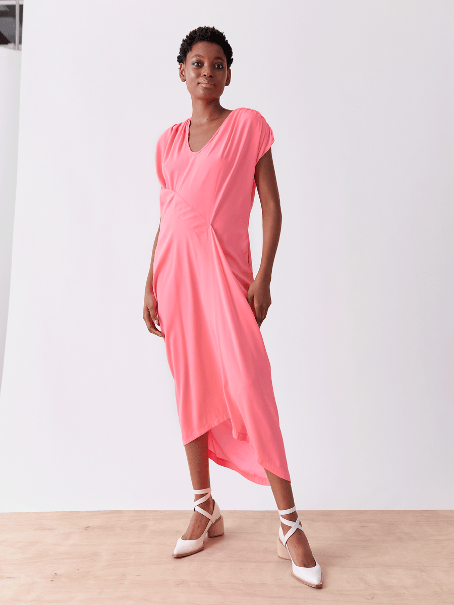 A person confidently wears the Long Ruched Fold Dress by Zero + Maria Cornejo, a bright pink silk charmeuse with short sleeves and an asymmetrical hem. They pair it with white pointed-toe shoes featuring crisscrossed straps, highlighted against a plain white background.