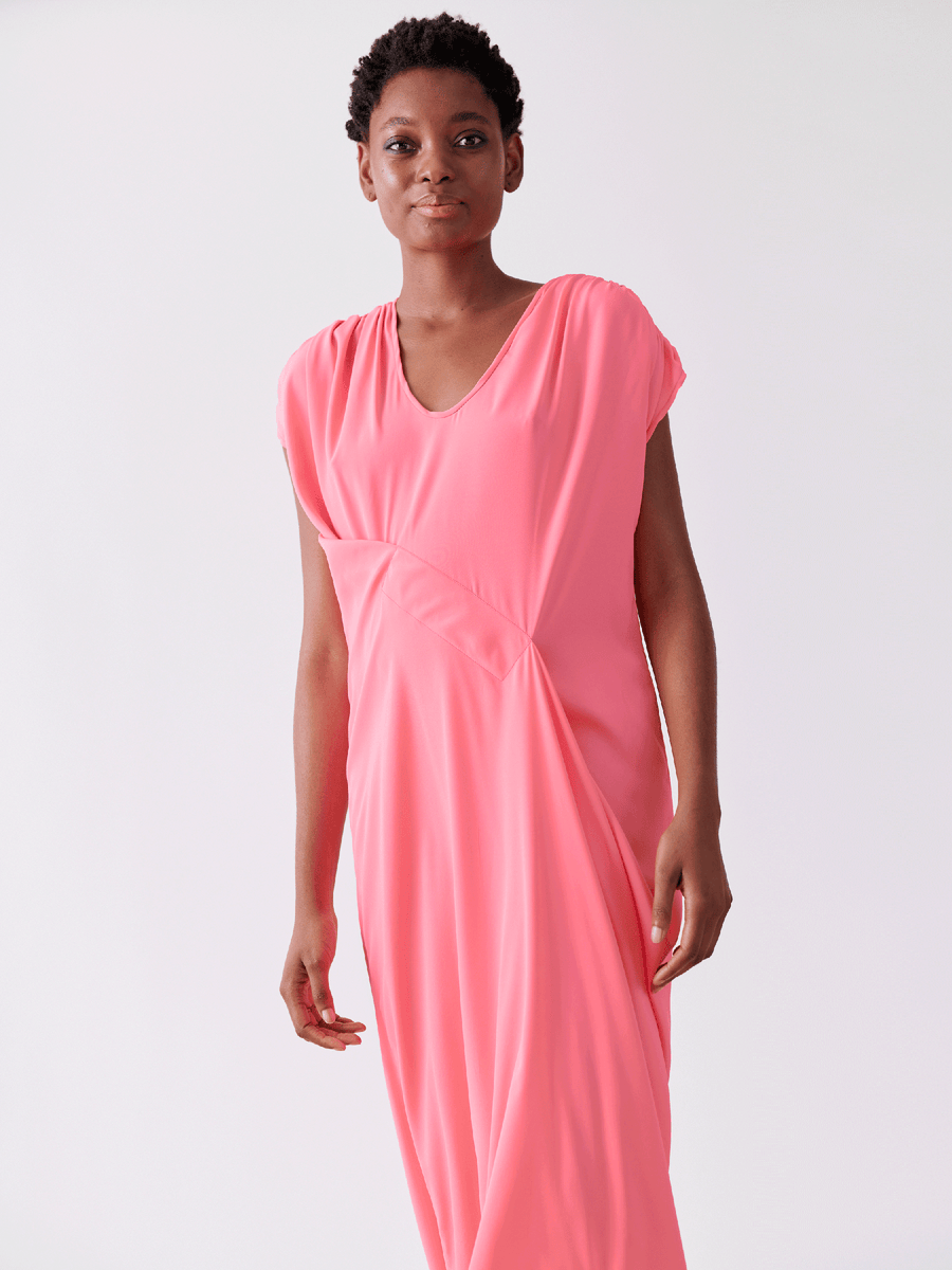 A person stands against a plain white background wearing the Long Ruched Fold Dress by Zero + Maria Cornejo, featuring pink silk charmeuse fabric and short sleeves, looking forward.