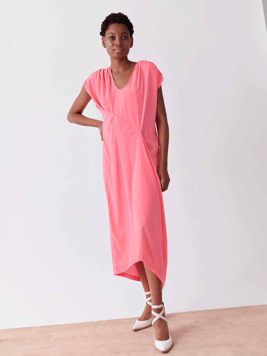 A person models the Long Ruched Fold Dress by Zero + Maria Cornejo in vibrant pink silk charmeuse, featuring a V-neck, short sleeves, and an asymmetrical design. They pair it with white crisscross-strap high heels against a minimalist wooden floor and light wall background.