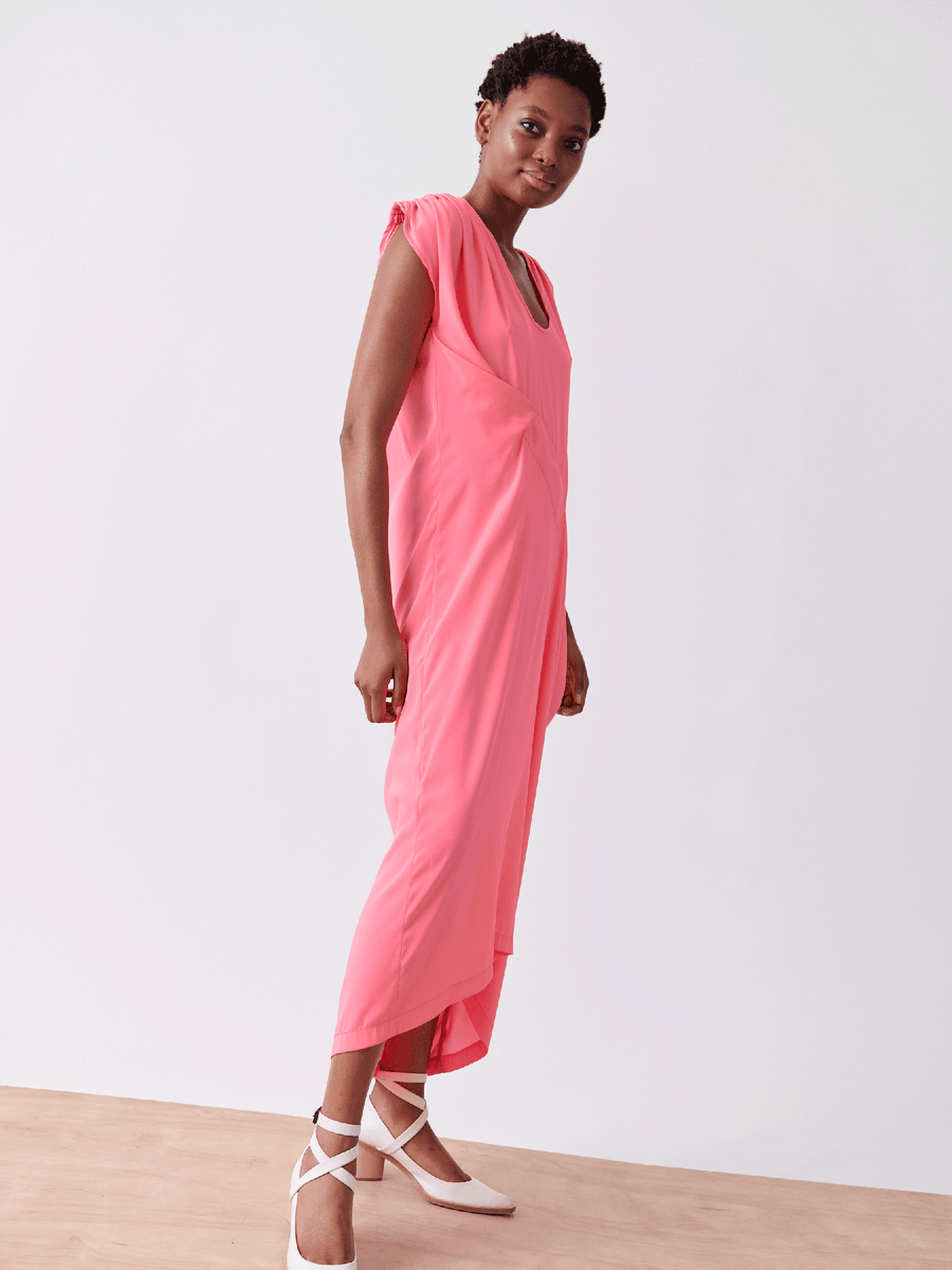 A person models the Long Ruched Fold Dress - Final Sale by Zero + Maria Cornejo, a flowing pink silk charmeuse piece with short sleeves. They stand against a white background on a wooden floor, donning white ballet flats with ankle straps and sporting short hair.