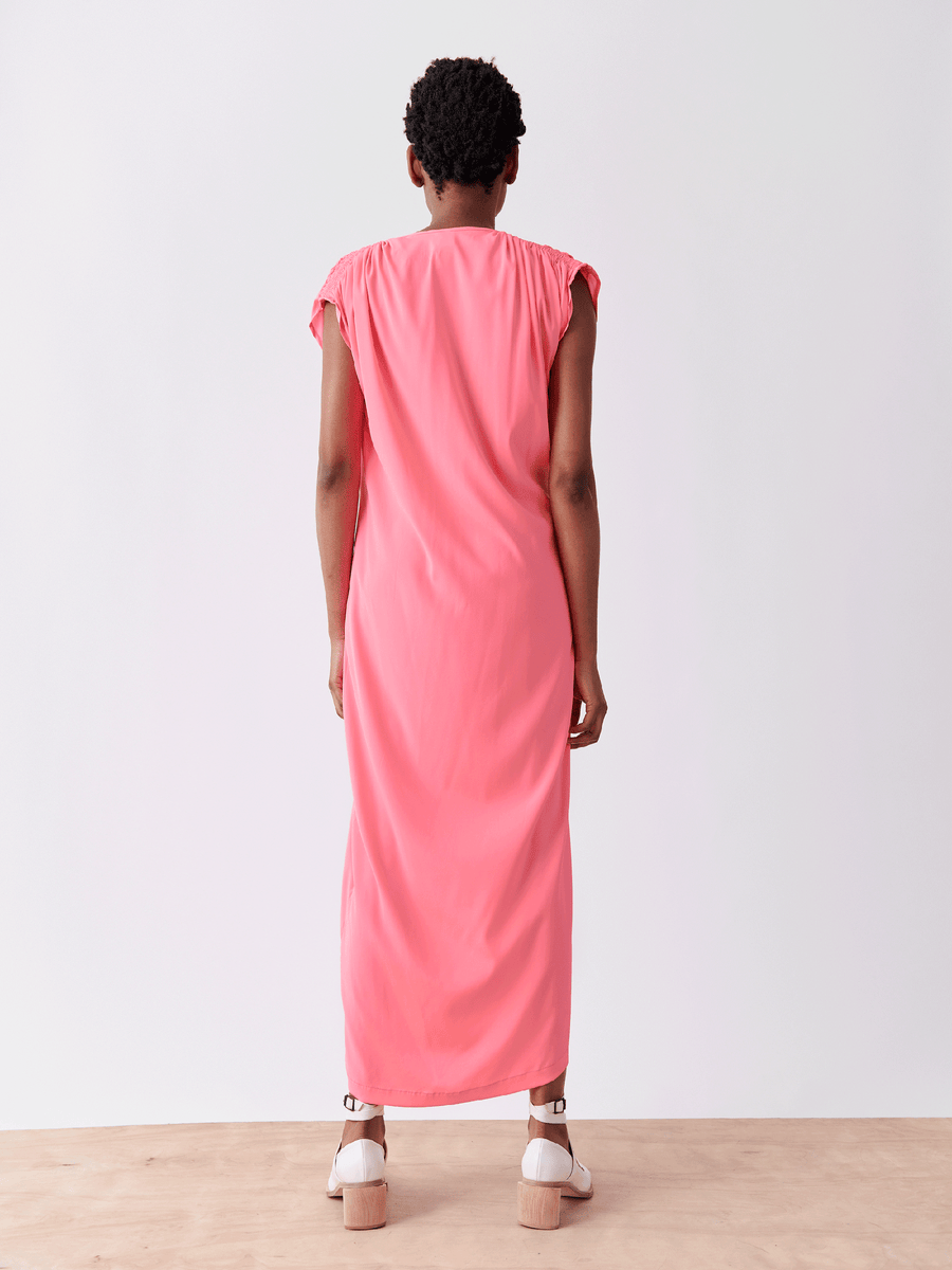 A person faces away from the camera, wearing Zero + Maria Cornejo's Long Ruched Fold Dress in bright pink silk charmeuse and white platform shoes with brown soles, against a plain, light background.