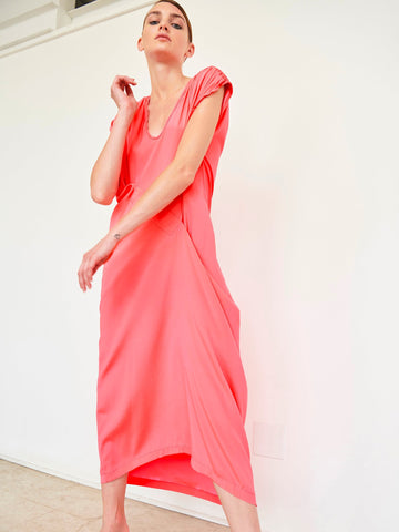 Someone poses in a Long Ruched Fold Dress by Zero + Maria Cornejo, against a white backdrop. This coral-pink silk charmeuse dress has short sleeves, an asymmetrical folded front, and a flowing mid-length skirt. They look down slightly with one arm bent, highlighting its elegant drape.