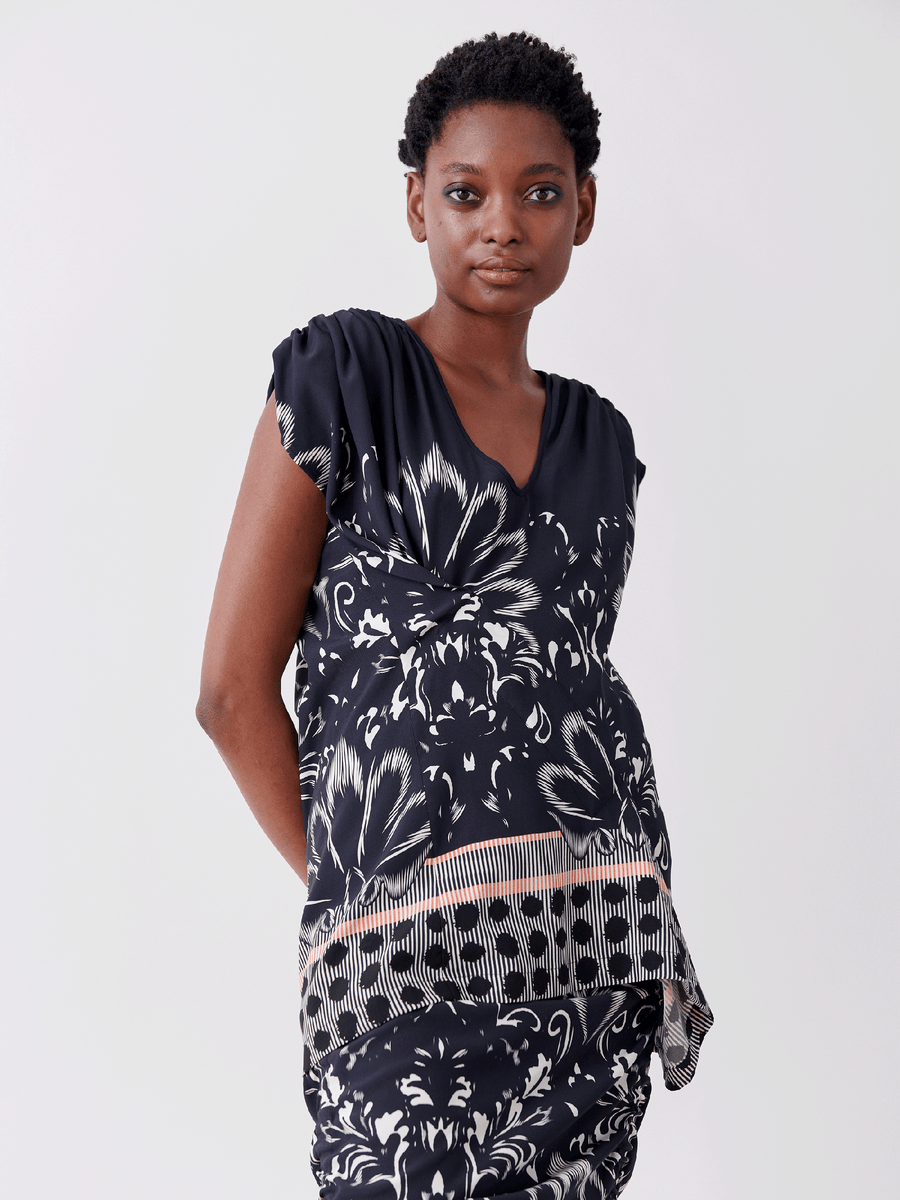 A person in a navy blue Zero + Maria Cornejo Ruched Fold Top, featuring white damask floral and geometric patterns, stands against a plain white background. They have short hair and a neutral expression, with their hands resting at their sides.