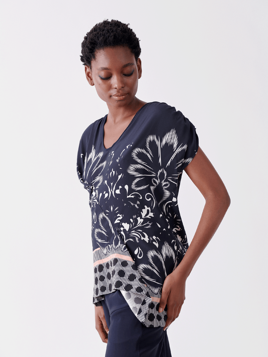 A person wearing the Ruched Fold Top by Zero + Maria Cornejo, featuring a navy blue silk charmeuse with a white damask floral pattern, glances down with one hand in their pocket against a plain white background.