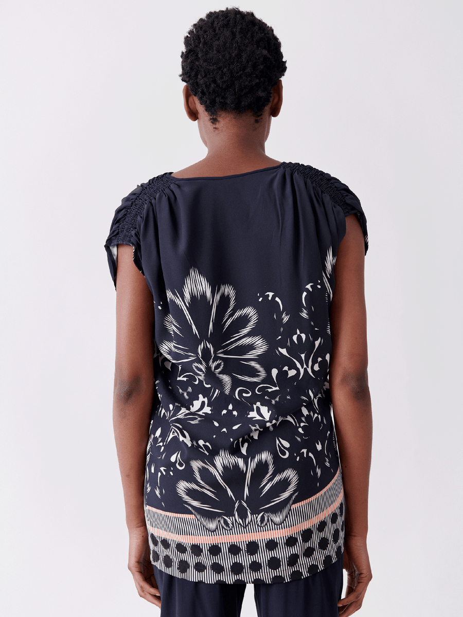 A person with short curly hair is facing away, wearing the Ruched Fold Top by Zero + Maria Cornejo. The dark blue silk top features white floral patterns, textured shoulders, and an orange and white decorative hem against a plain light gray background.