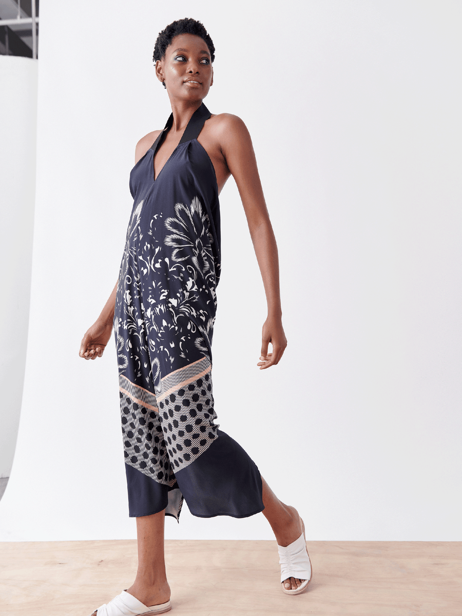 A person confidently walks barefoot on a light wooden floor in a sleeveless Long Gigi Dress by Zero + Maria Cornejo, crafted from navy blue stretch charmeuse with floral and geometric patterns; all set against a plain white background.