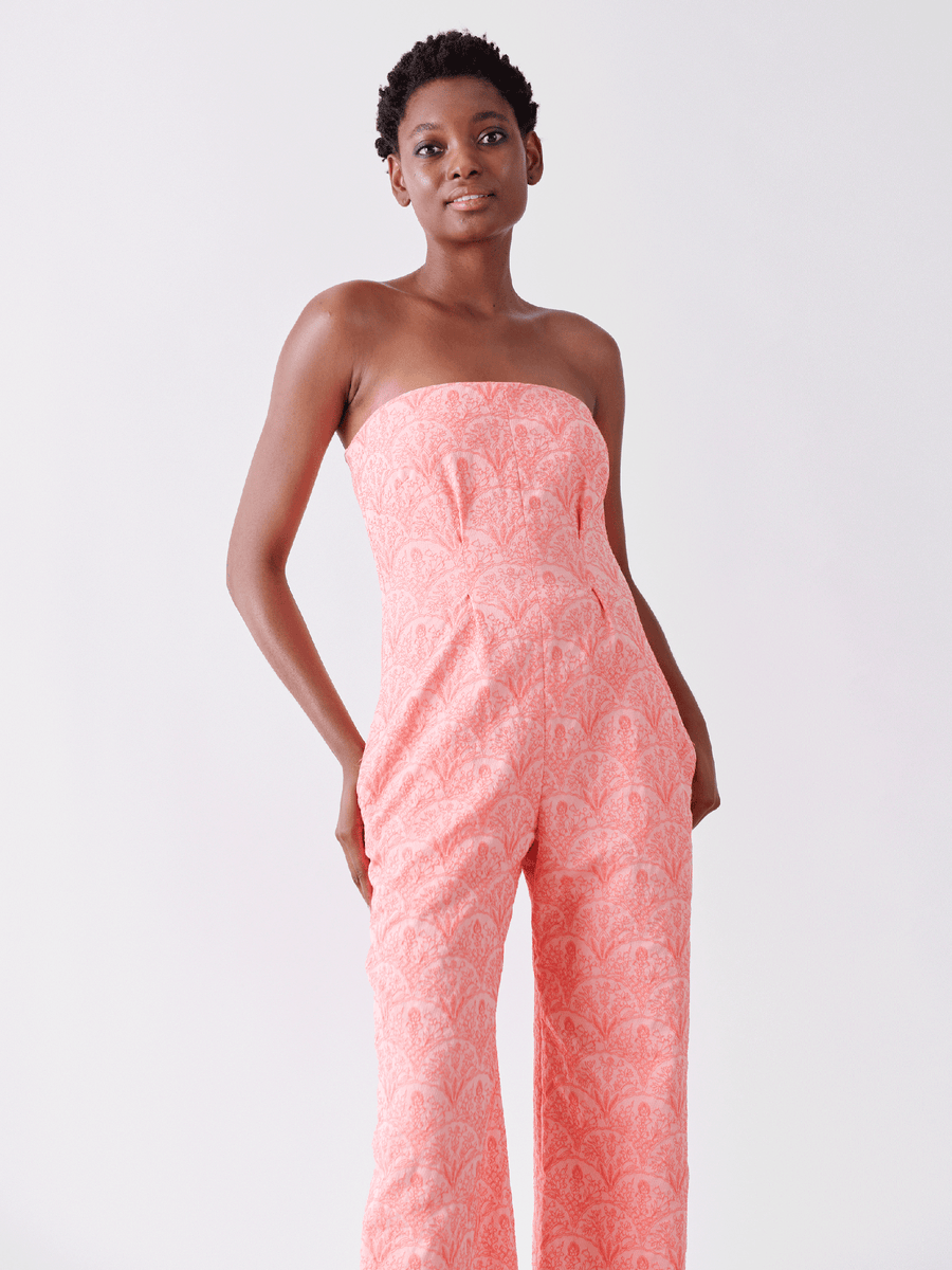 A person in the Pia Jumpsuit by Zero + Maria Cornejo, featuring a strapless design made from recycled polyester with a subtle pattern, stands against a plain white background. They have short hair, are smiling, and have their hands relaxed by their sides.