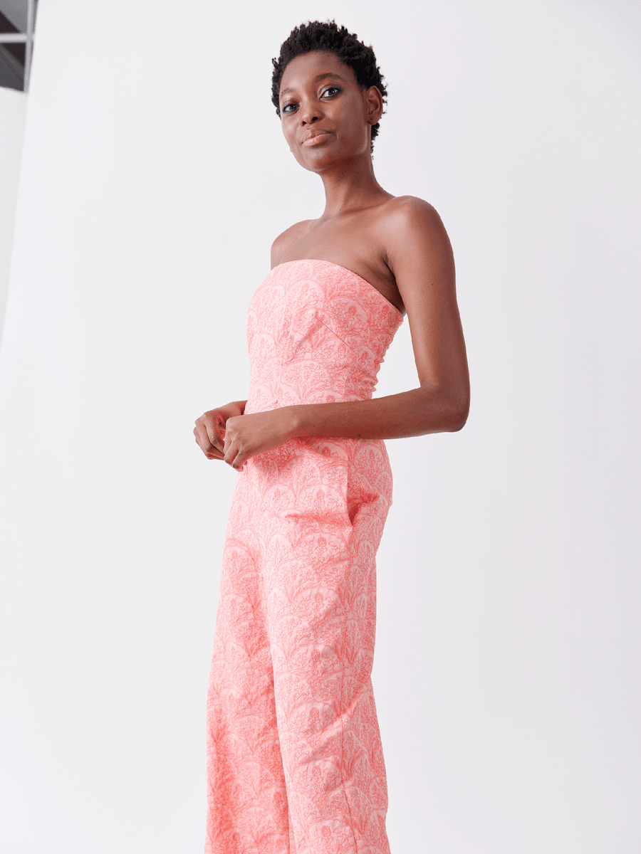 A person modeling the Pia Jumpsuit - Final Sale by Zero + Maria Cornejo, made from recycled polyester and featuring a strapless pink pattern, stands confidently against a plain white background with short hair, gazing slightly away from the camera.