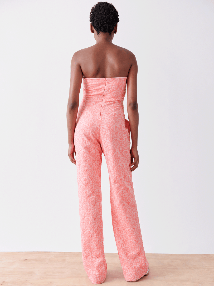 Rear view of a person with short hair wearing the Pia Jumpsuit by Zero + Maria Cornejo in jacquard coral pink, standing on a light wood floor against a plain white background.