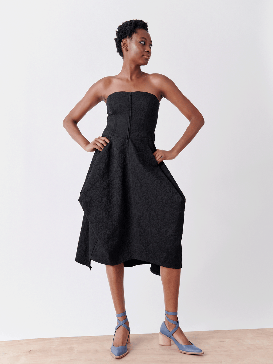 Against a white backdrop, a person exudes confidence in the Zero + Maria Cornejo's black Strapless Nio Dress with a flared skirt made from recycled polyester. With short black hair and blue heeled sandals, they pose with hands on hips, gazing aside.