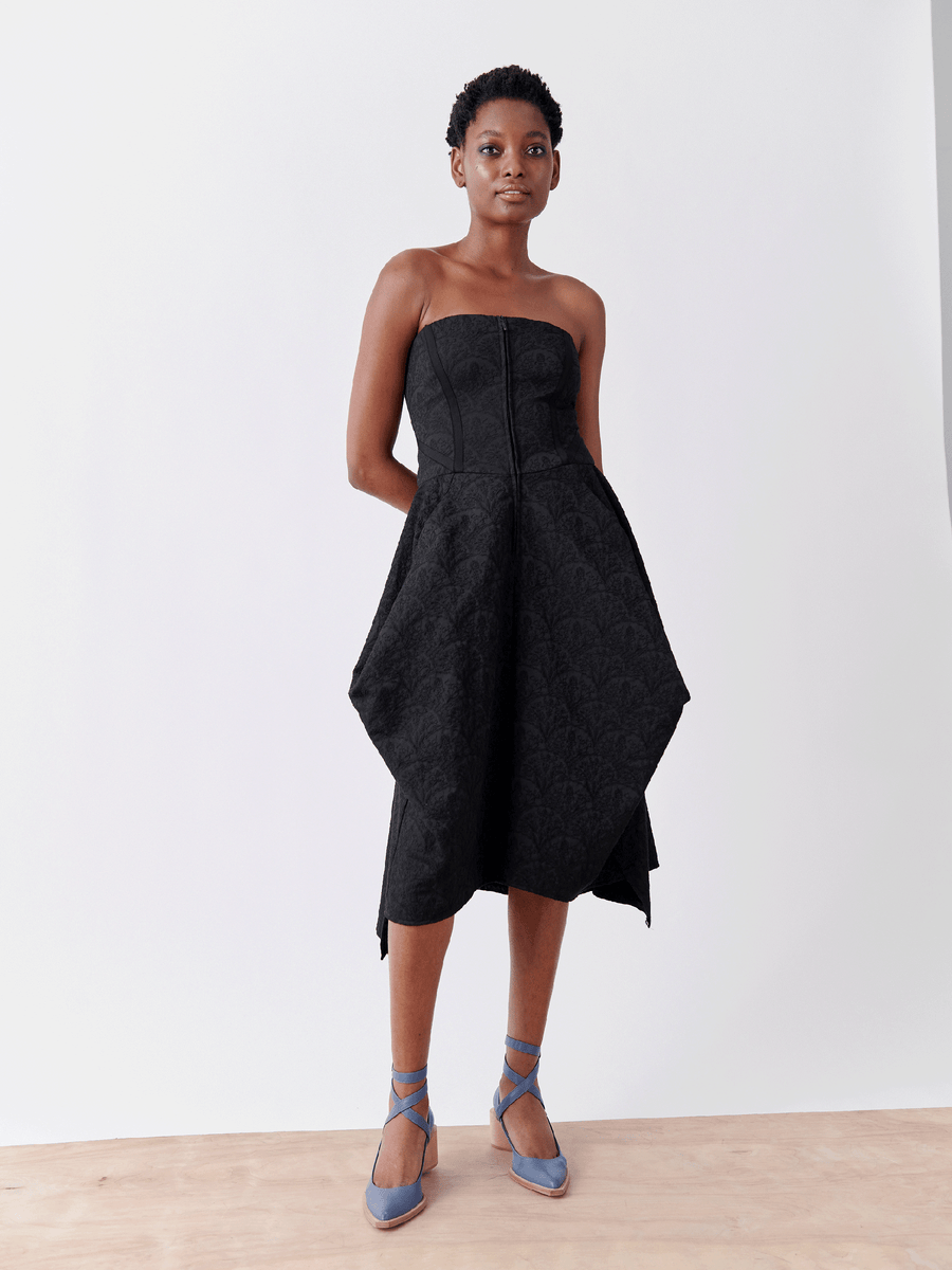 A person with short hair stands confidently against a plain background, wearing the Strapless Nio Dress by Zero + Maria Cornejo, made from recycled polyester. Blue strappy heels accentuate the elegant hemline and their poised, relaxed posture.