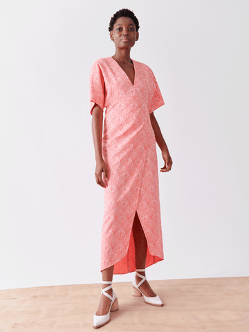 Against a plain background, a person wears the Wrap Eri Dress by Zero + Maria Cornejo, featuring pink floral patterns, short sleeves, and a V-neckline. Made from recycled polyester with a front slit, the look is completed with white block-heel strappy shoes.