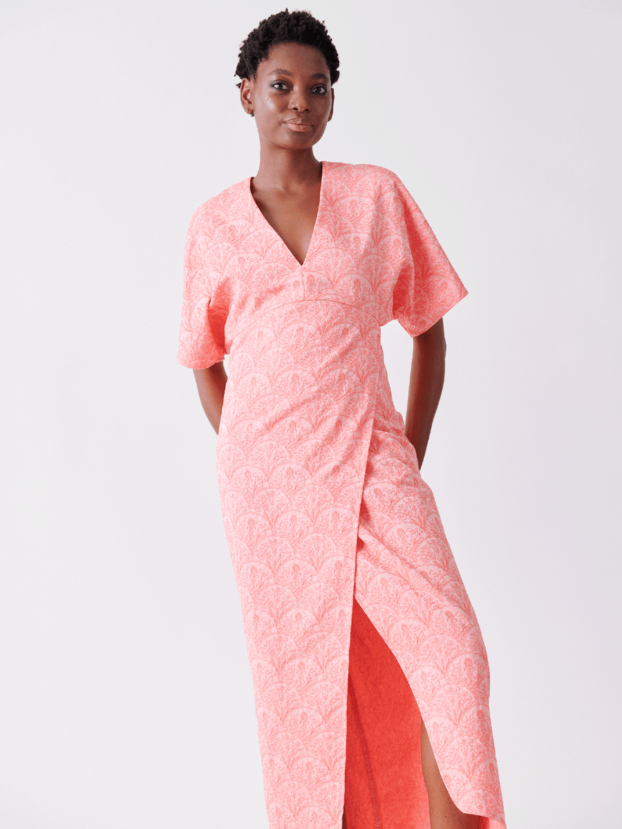 A person confidently poses with short hair and a relaxed stance, hands behind their back, against a light background. They're wearing the Zero + Maria Cornejo Wrap Eri Dress in pink floral recycled polyester, featuring short sleeves and a slit.