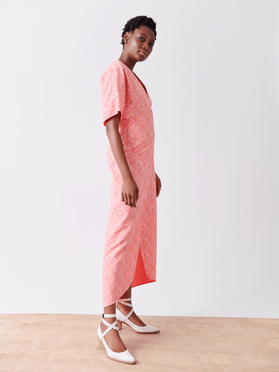 A person stands against a plain backdrop wearing the Wrap Eri Dress by Zero + Maria Cornejo, featuring a pink floral pattern and crafted from recycled polyester. They complement it with white heels, exhibit a neutral expression, and stand on a wooden floor in a minimal setting.