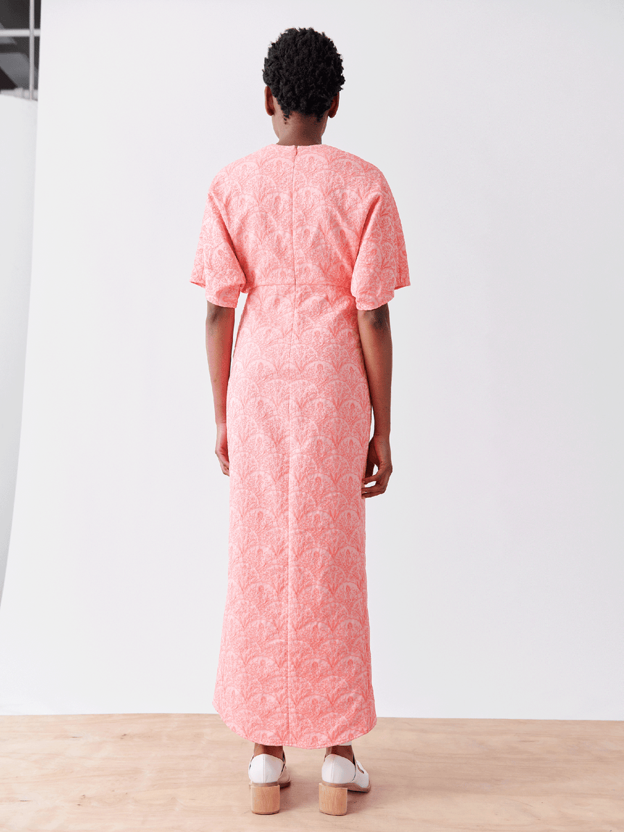 A person in a coral Wrap Eri Dress by Zero + Maria Cornejo, featuring a floral stretch jacquard design, stands on a wooden floor with white shoes against a light gray backdrop.