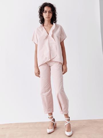 Against a white backdrop, an individual donning a light pink blouse pairs it with Zero + Maria Cornejo's Slim Flight Pant, made from lightweight tech fabric. Their curly hair complements the minimalist, stylish appearance completed with white ballet flats featuring ribbons.