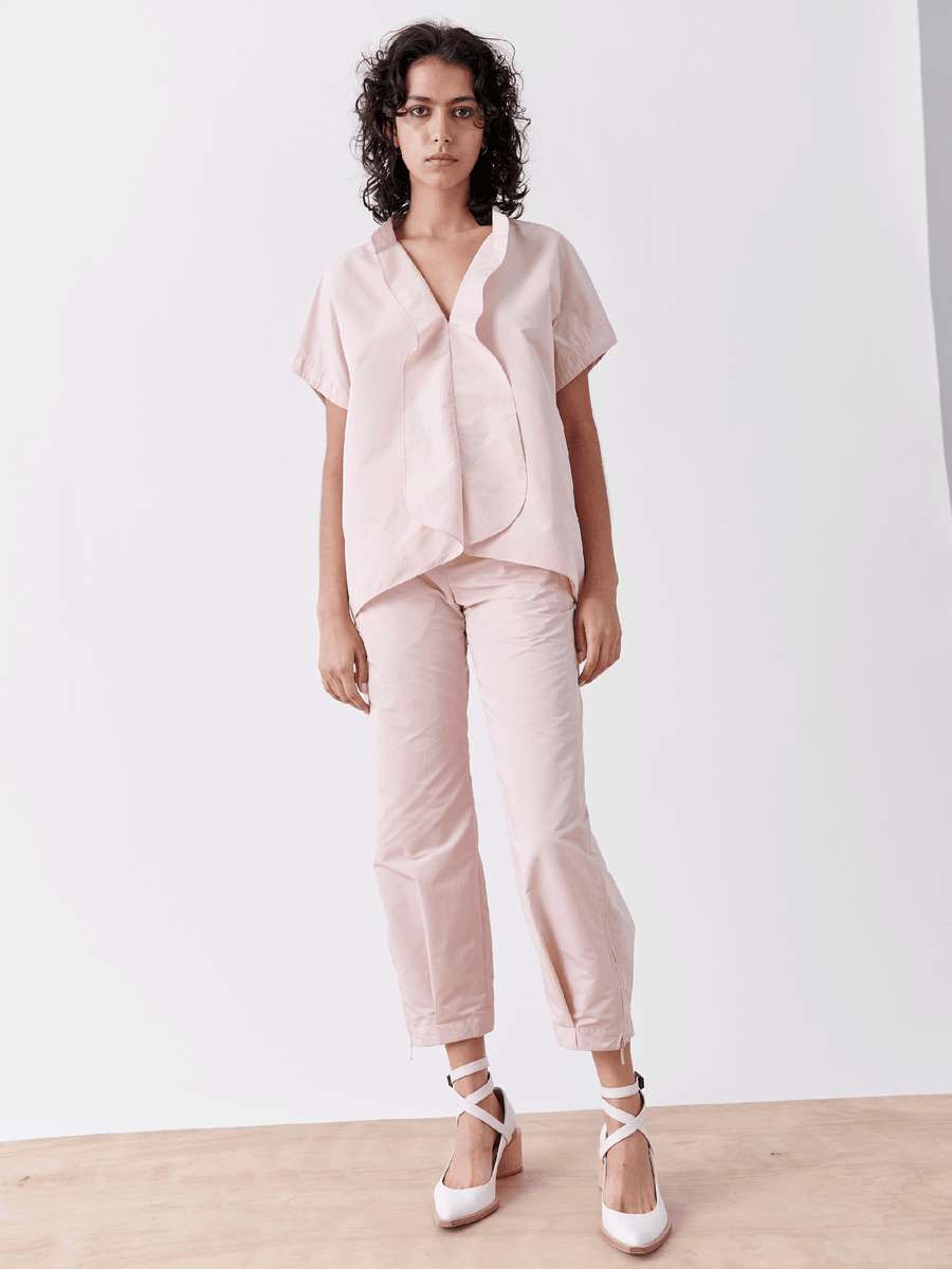 Against a white backdrop, an individual donning a light pink blouse pairs it with Zero + Maria Cornejo's Slim Flight Pant, made from lightweight tech fabric. Their curly hair complements the minimalist, stylish appearance completed with white ballet flats featuring ribbons.