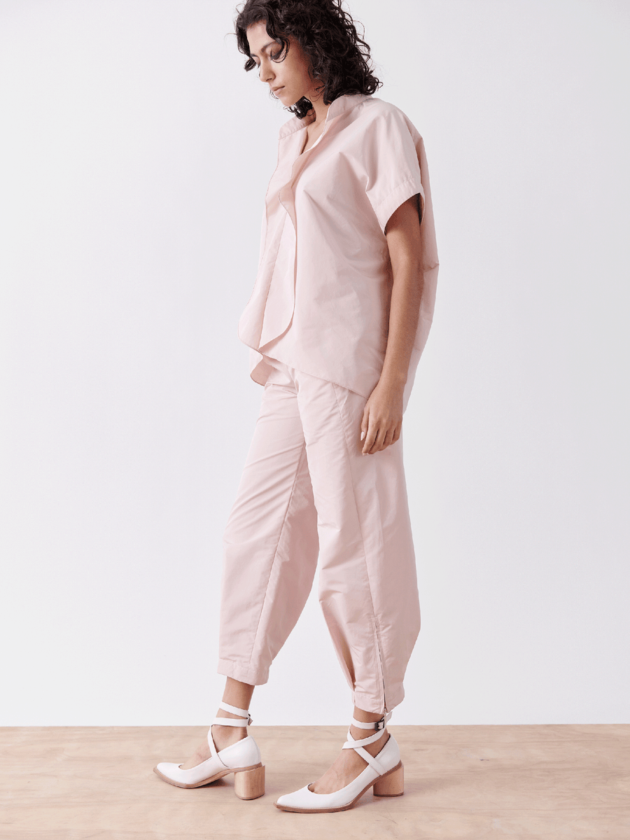 Against a plain white background, a curly-haired person wears the Zero + Maria Cornejo Slim Flight Pant in light pink, made from lightweight tech fabric, paired with white, closed-toe block heels. They maintain a relaxed side-facing posture.