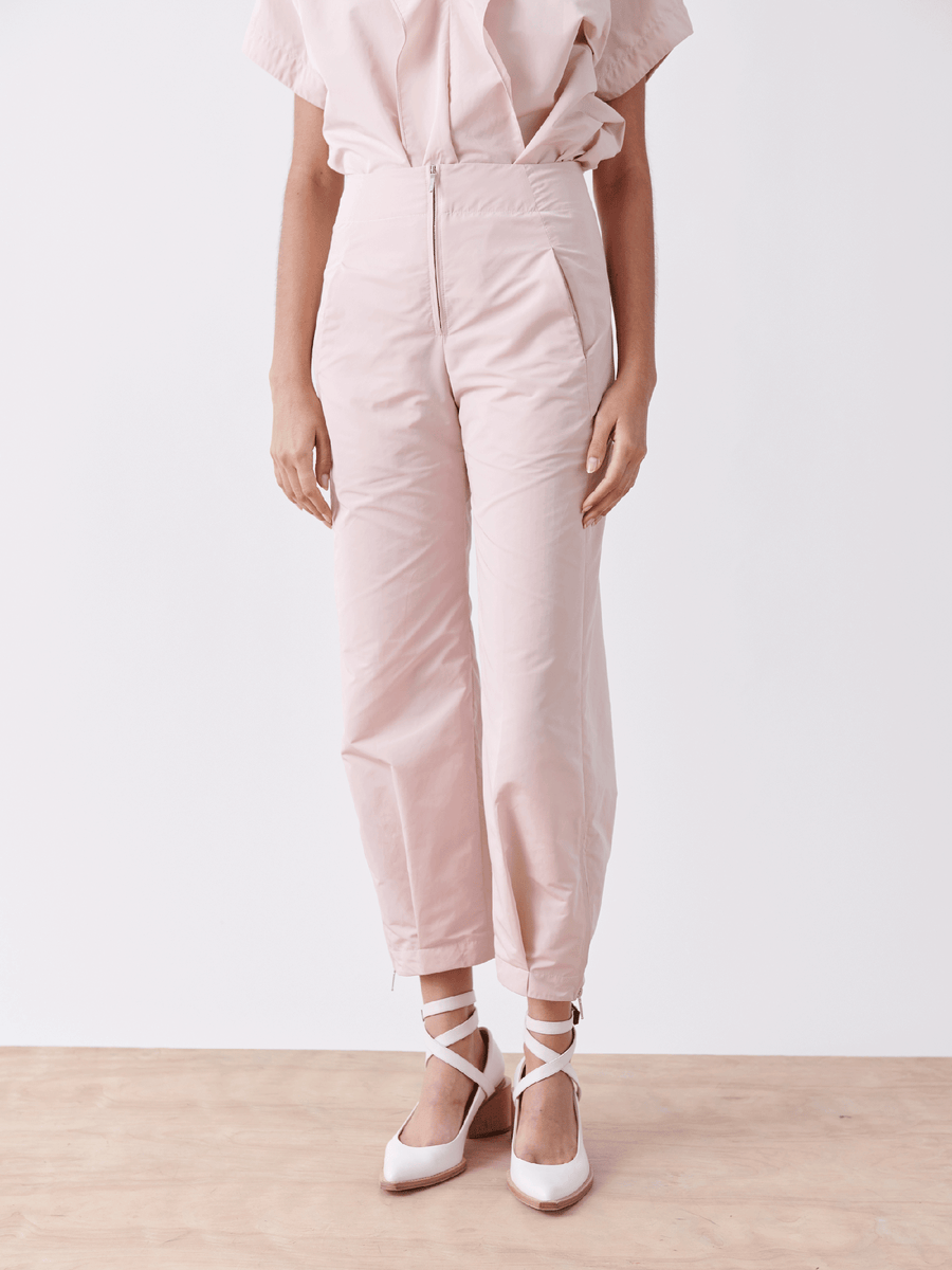 A person wearing Zero + Maria Cornejo's Slim Flight Pant in light pink with a matching blouse stands on a wooden floor. The high-waisted, technical woven pants are paired with white ankle-strap shoes, creating a soft and elegant look against a plain white background.