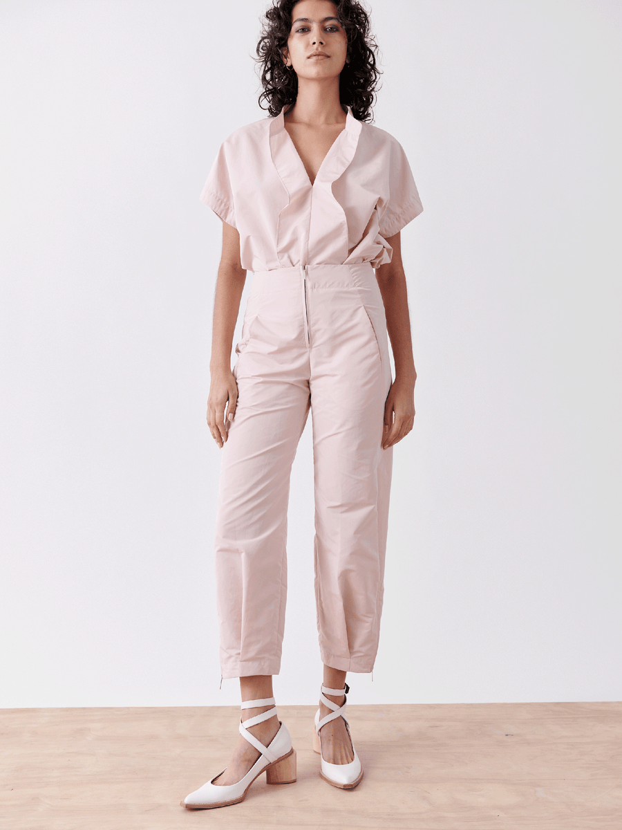 A person wears a light pink short-sleeve shirt, Zero + Maria Cornejo's Slim Flight Pant - Final Sale, and white ankle-strap shoes. They stand on a wood floor against a plain white background.