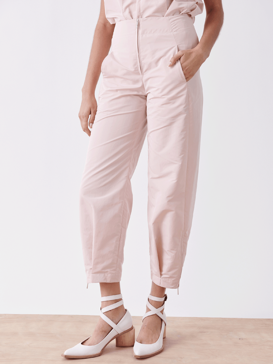 A person is wearing Zero + Maria Cornejo's Slim Flight Pant in pale pink with a front zipper and ankle zippers, made from lightweight tech fabric, with a hand in one pocket. They are also wearing white strappy block-heeled shoes against a plain background.