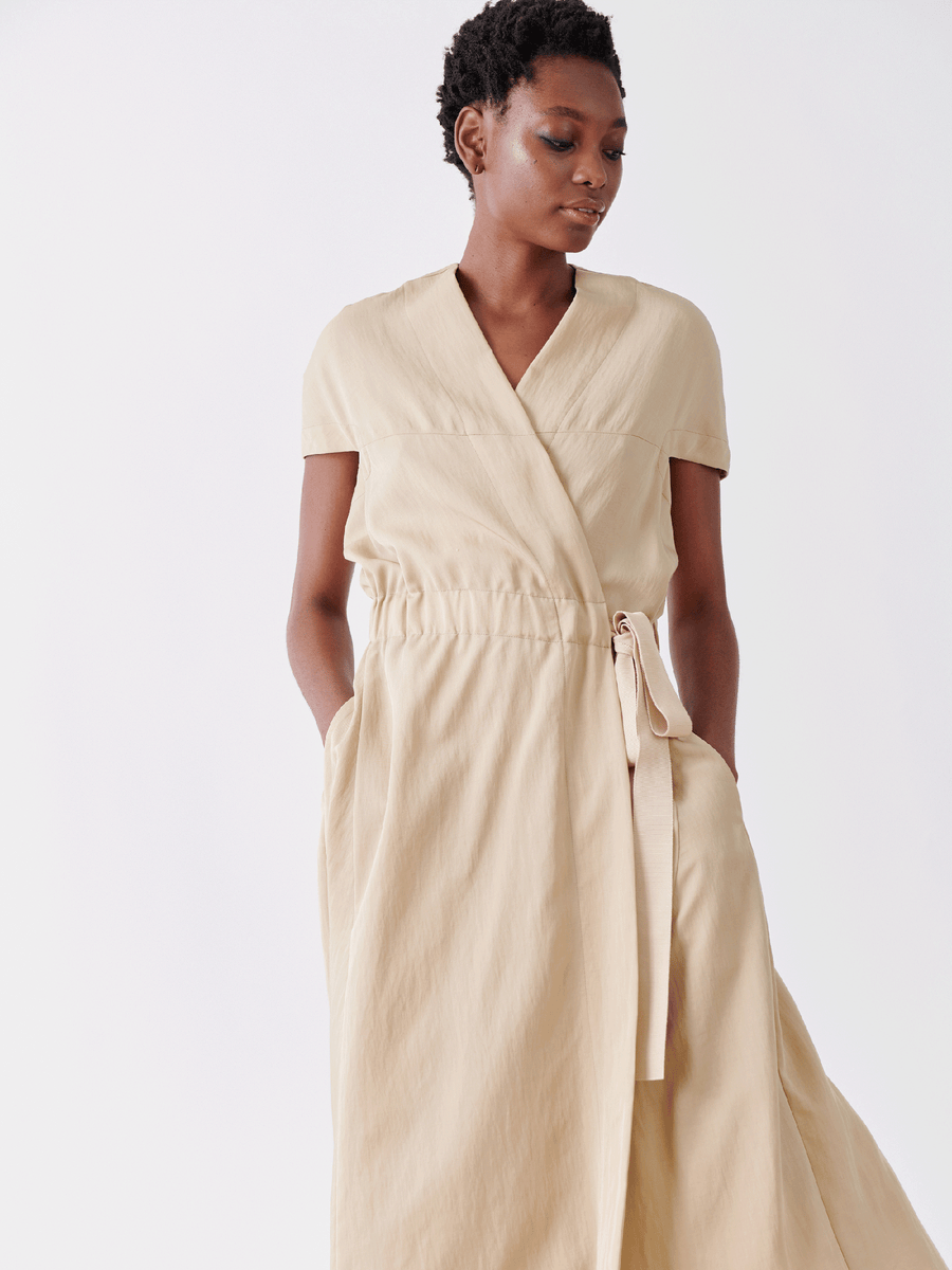 A model poses against a plain white background, looking down to the side in Zero + Maria Cornejo's Wrap Hex Dress - Final Sale. The light beige short-sleeved dress features textured recycled viscose fabric and an asymmetrical hem, with the model's hands in the pockets.