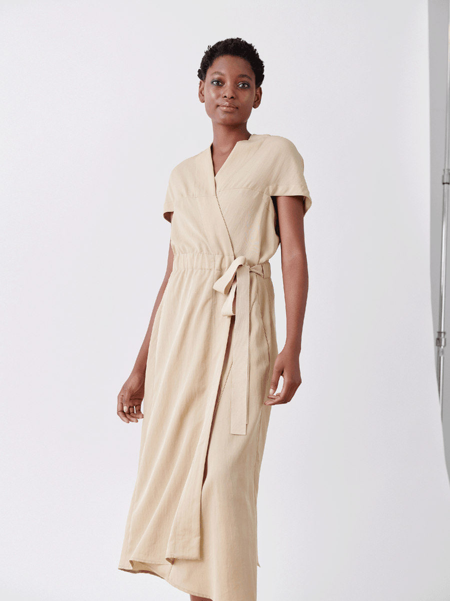 A person stands against a white backdrop, wearing Zero + Maria Cornejo's beige Wrap Hex Dress made from recycled viscose. Tied at the waist with a matching belt and featuring an asymmetrical hem, their short hair and calm expression exude effortless style.