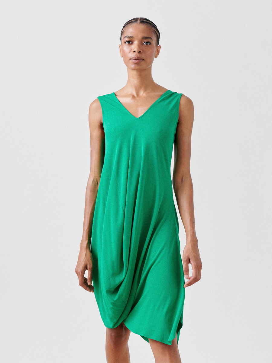 A person with short hair wears the Zero + Maria Cornejo Opposite Dress—Final Sale, a sleeveless, draped green V-neck dress made from lightweight fabric. Featuring front gathers and an asymmetrical skirt, they stand against a plain white background.