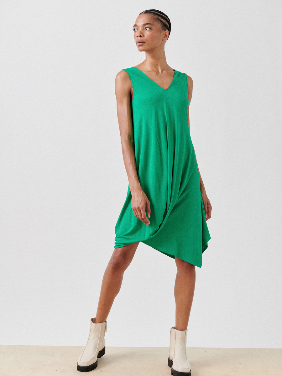 The person wears the Opposite Dress by Zero + Maria Cornejo, a sleeveless green piece with an asymmetrical hem made from lightweight fabric. Paired with light-colored ankle boots, they confidently pose against a plain white background, one hand on their hip.