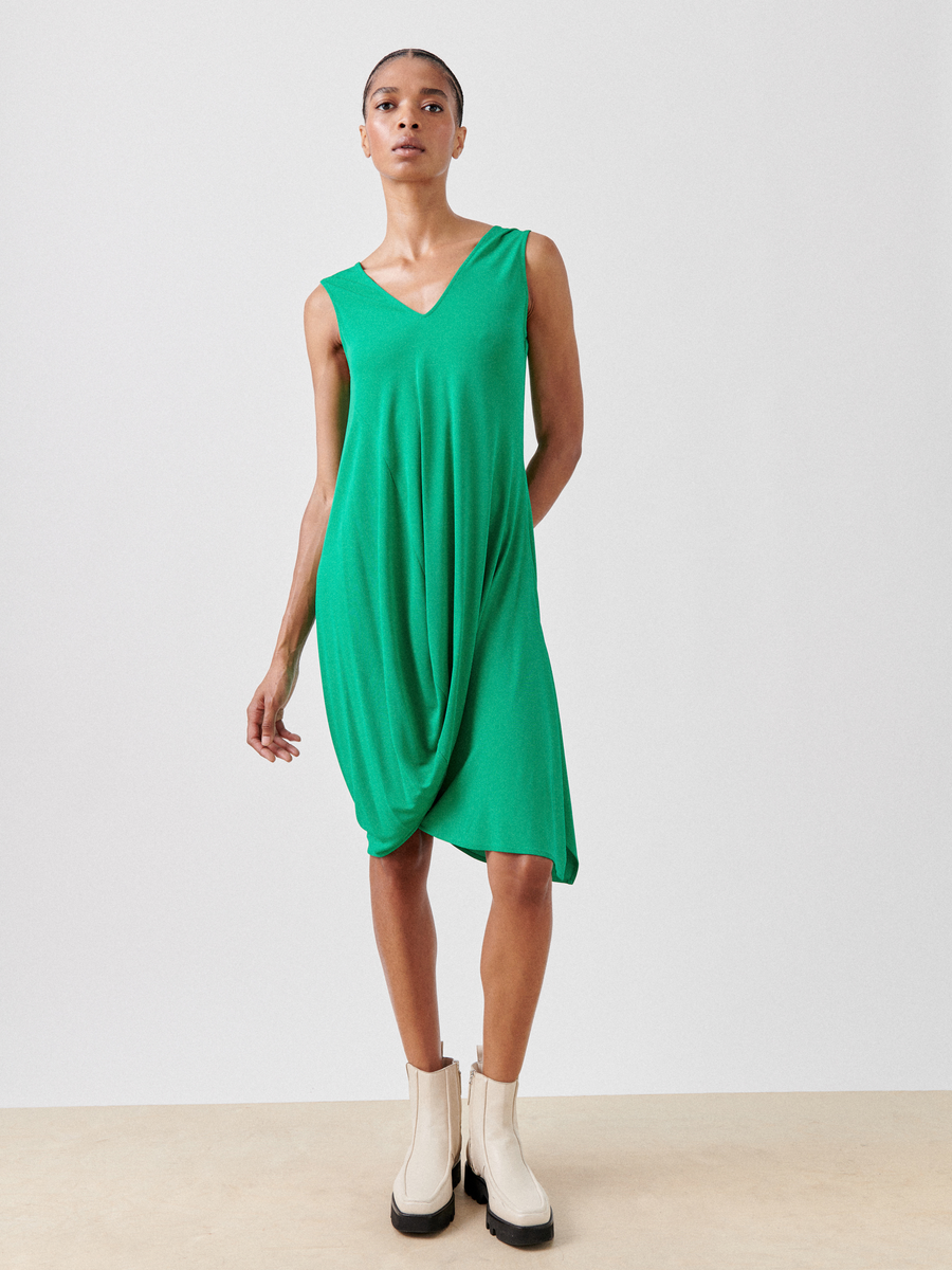 A person in the Zero + Maria Cornejo Opposite Dress - Final Sale, a sleeveless green draped design made from lightweight fabric, stands against a plain background. They're wearing beige ankle boots and face forward with their arms relaxed by their sides.