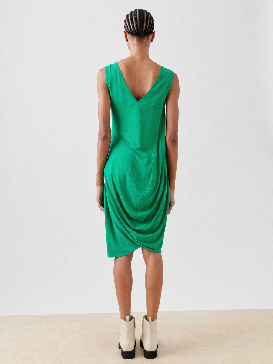 A person is seen facing away in the Opposite Dress by Zero + Maria Cornejo, a sleeveless green dress made from lightweight fabric with a draped back. Their braided hair pairs perfectly with light-colored ankle boots against a plain, light background.