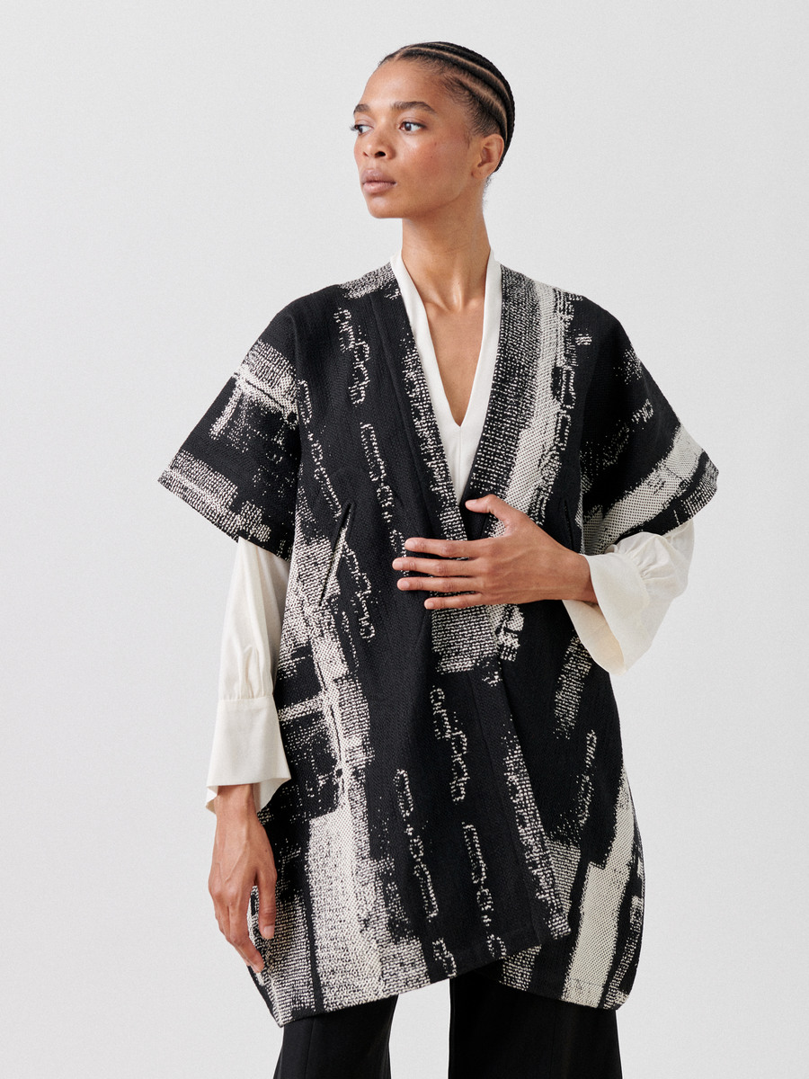 A person with short braided hair wears a Gaban Coat by Zero + Maria Cornejo, featuring a black and white jacquard pattern, over a long-sleeved white blouse. They pose against a plain background with one hand resting on their chest.