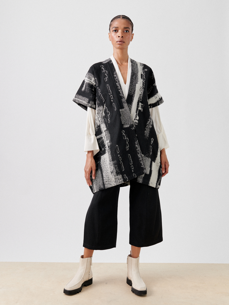 Dressed in a loose-fitting Gaban Coat by Zero + Maria Cornejo with wide sleeves over a white shirt, and black culottes paired with light-colored ankle boots, they exude confidence against a plain background. Made from recycled fibers, this stylish ensemble is part of the Final Sale collection.