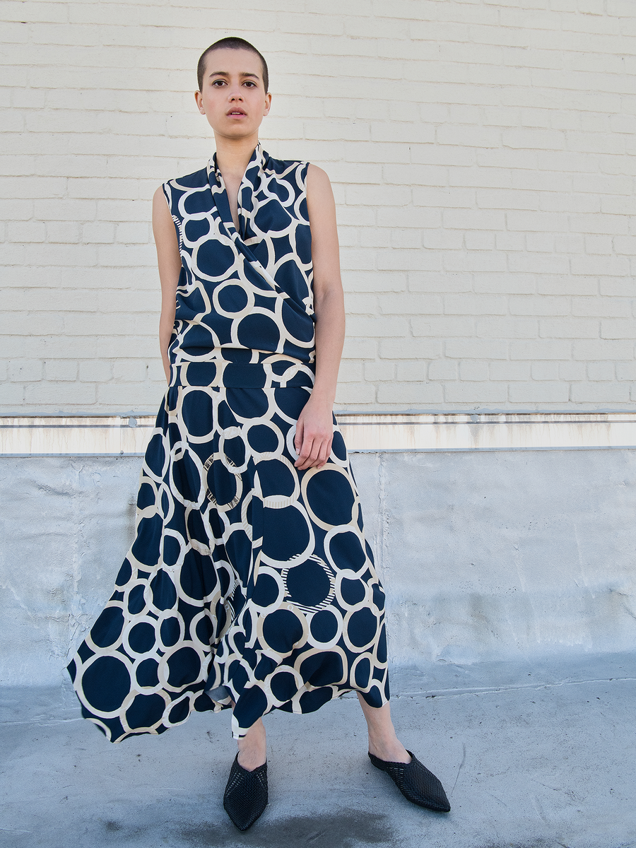 Against a brick wall, a person wears Zero + Maria Cornejo's Circle Skirt - Final Sale, featuring a bold black-and-white circle pattern. With short hair and black pointed shoes, they pose outdoors on concrete.