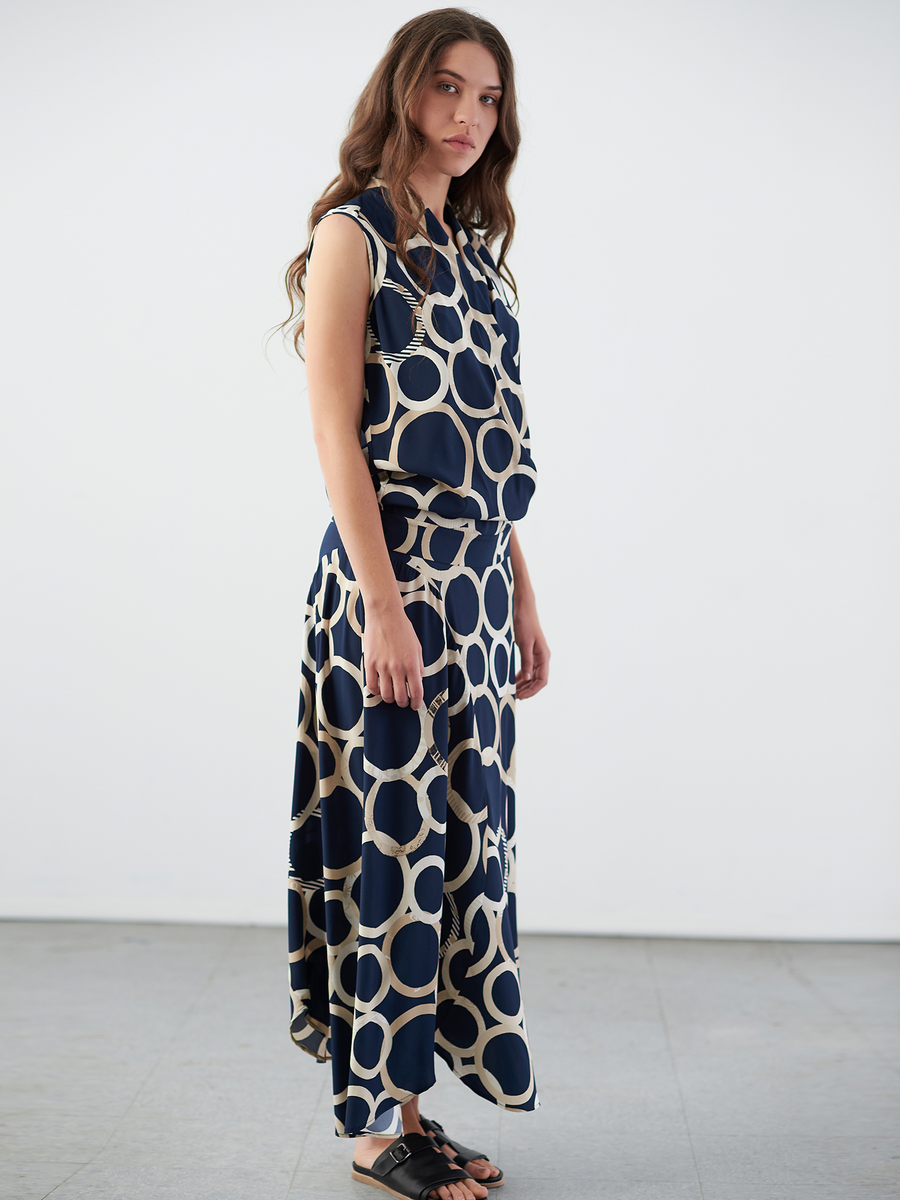 A woman with long brown hair in a navy blue dress adorned with large white circles, stands against a plain background. She looks to her left, hands relaxed at her sides, while wearing black sandals and the chic Circle Skirt by Zero + Maria Cornejo.