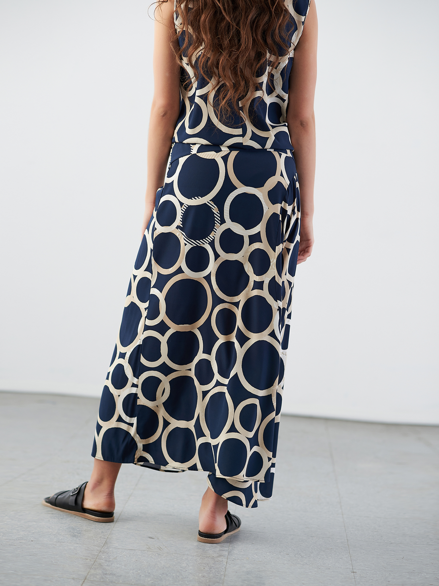 A person with long, wavy hair stands facing away on a light gray floor, wearing the Circle Skirt by Zero + Maria Cornejo. The skirt is navy blue and beige with large circle patterns, paired with black slip-on shoes.