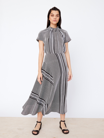 A woman stands against a white background wearing Zero + Maria Cornejo's Circle Skirt - Final Sale dress, featuring vertical and diagonal black-and-white stripes, a banded waist, short sleeves, and a flared textured skirt. Her long dark hair accompanies her look with black strappy sandals.