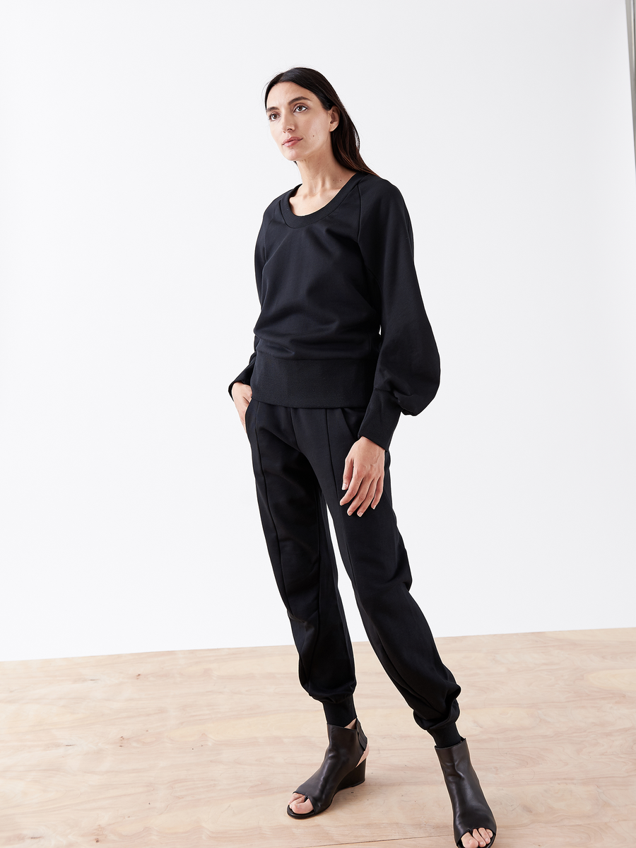 On a wooden floor, a person with long dark hair wears a black long-sleeve top, Zero + Maria Cornejo Akeo Sweatpant - Final Sale in cotton terrycloth with an elastic banded waist, and black open-toed shoes against a plain white background.