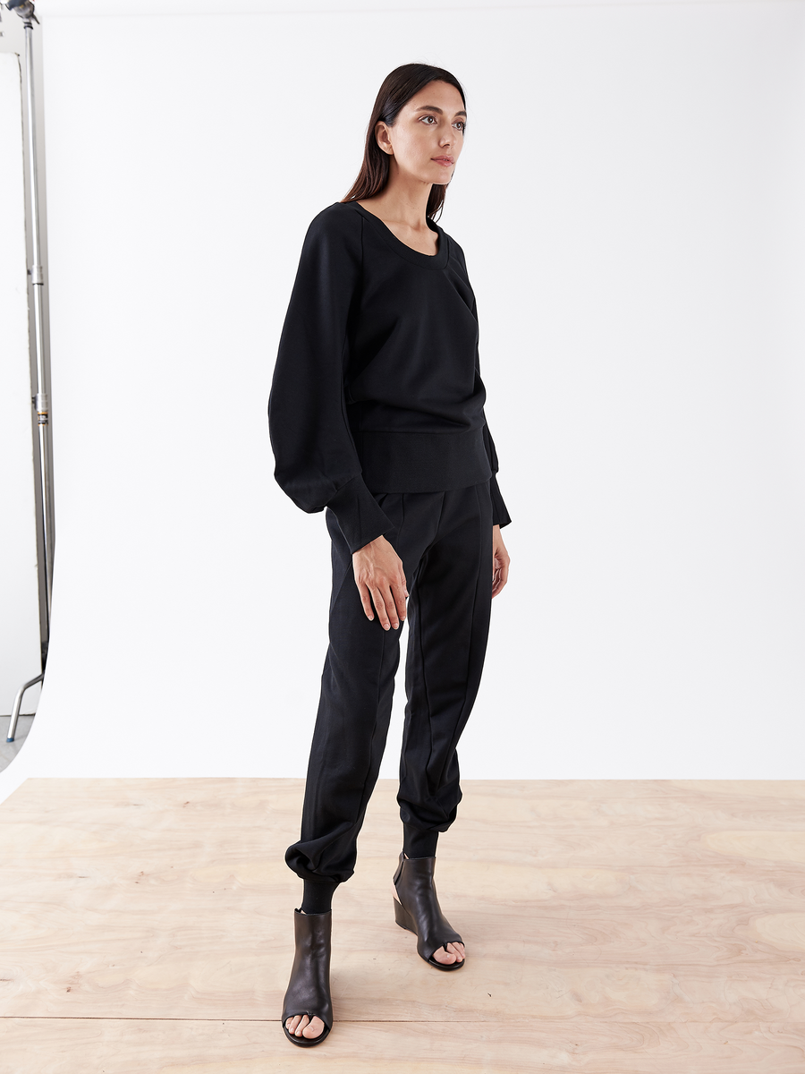 A woman stands on a wooden floor against a white background, wearing a black long-sleeve top and Zero + Maria Cornejo's Akeo Sweatpant with an elastic waist, paired with open-toed black ankle boots. She gazes to the side.