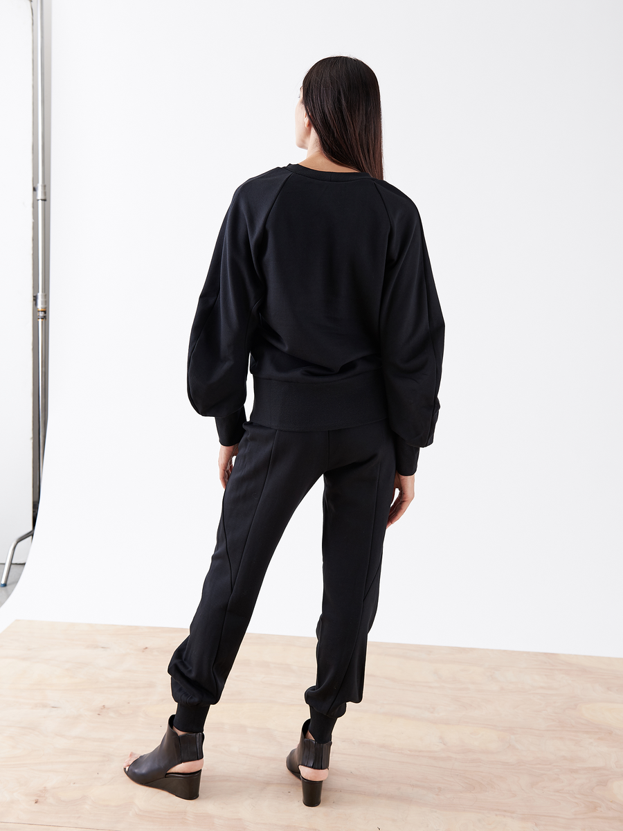 A woman with long dark hair stands on a wooden floor, facing away. She wears a black French Terry sweatshirt, Zero + Maria Cornejo's Akeo Sweatpant - Final Sale with an elastic-banded waist, and black wedge-heeled shoes. The plain white wall serves as a minimalist backdrop.