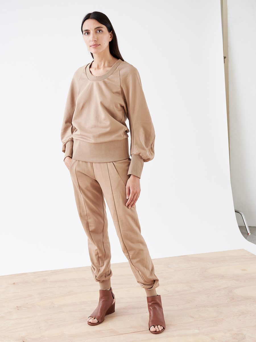 A woman wears a beige French Terry long-sleeve top and Zero + Maria Cornejo's Akeo Sweatpant in cotton terrycloth, standing on a wooden floor against a white backdrop. She pairs the outfit with brown open-toe heels, her long dark hair framing her face as she gazes neutrally at the camera.