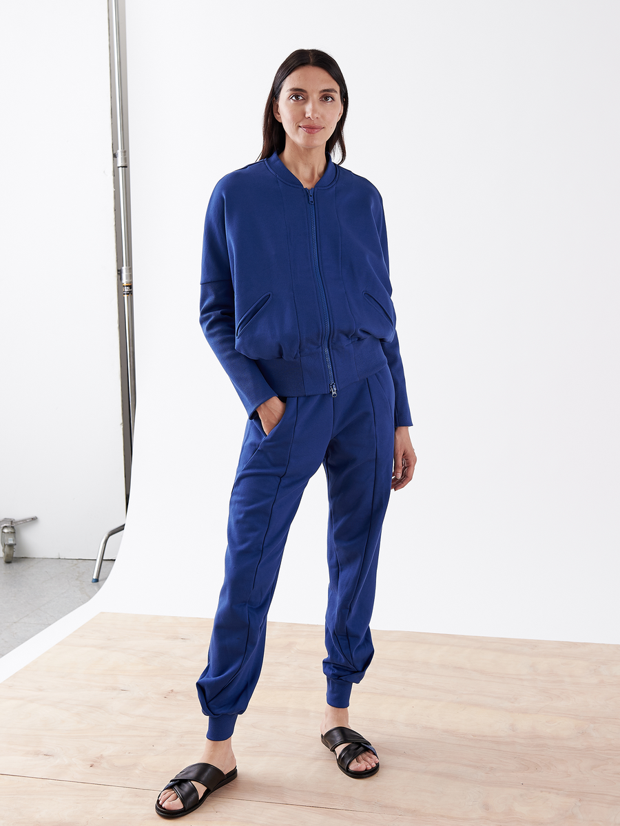 A person in a matching blue Akeo Sweatpant - Final Sale by Zero + Maria Cornejo with an elastic-banded waist and black sandals stands on a wooden floor against a plain white background, hands in pockets, with long dark hair.