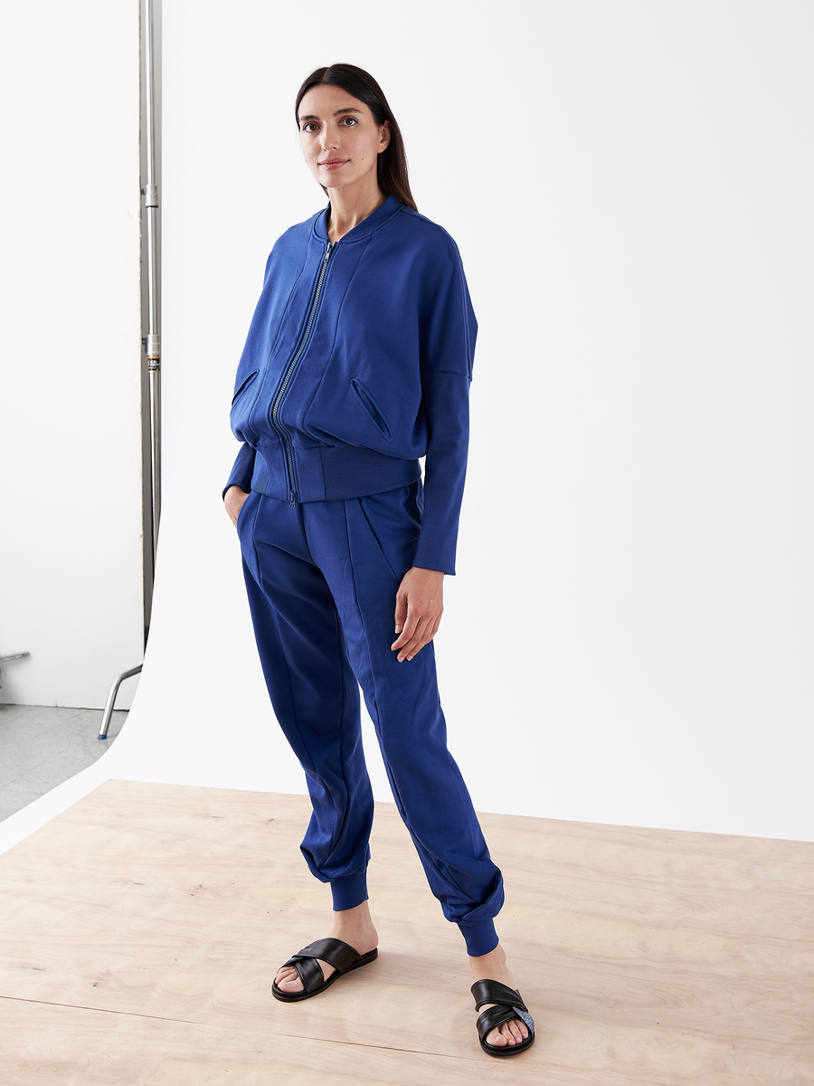 A person stands on a wooden floor, wearing the Akeo Sweatpant from Zero + Maria Cornejo—a blue zip-up jacket and black sandals. The background is plain white with a metal stand partially visible.