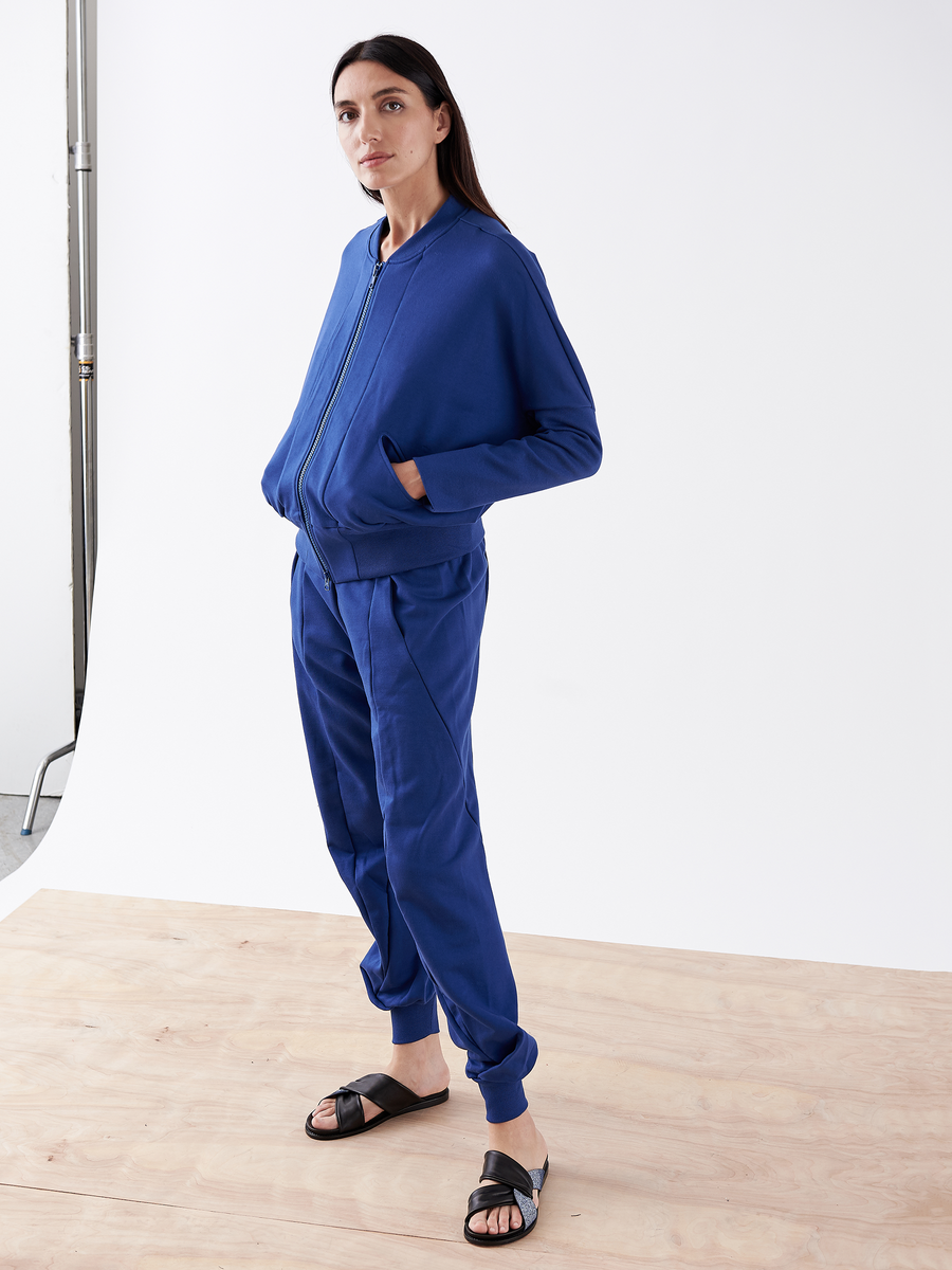 A person with long dark hair stands on a wooden floor, wearing Zero + Maria Cornejo Akeo Sweatpants in blue and black sandals. Set against a plain white background, they gaze at the camera with hands in elastic-banded waist pockets.