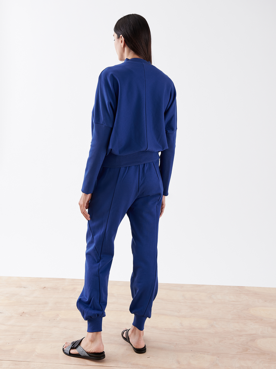 A person with long dark hair stands on a wooden floor, wearing the Akeo Sweatpant from Zero + Maria Cornejo in blue French Terry with an elastic waist and black sandals. They face away, highlighting the outfit against a plain white background.