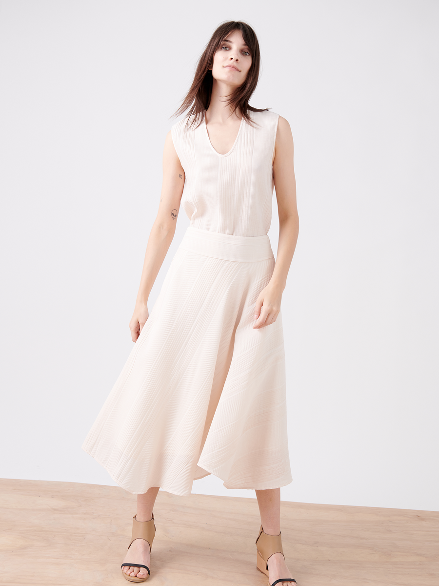 A woman stands on a wooden floor wearing the Aki Wave Skirt by Zero + Maria Cornejo. The sleeveless, cream-colored dress features an asymmetrical skirt and banded waist. With her long dark hair flowing as she looks forward, she completes the look with beige sandals against a plain white backdrop.