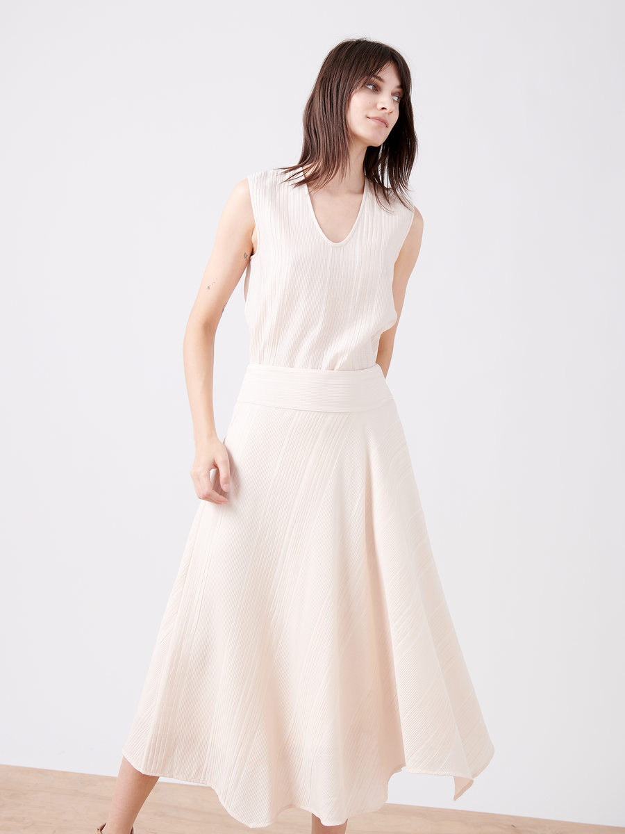 A woman stands against a white backdrop, wearing the sleeveless Aki Wave Skirt of Zero + Maria Cornejo in cream with a V-neckline and an asymmetrical design. Her shoulder-length brown hair complements her relaxed sideward gaze, while the dress’s elegant sateen finish adds sophistication.