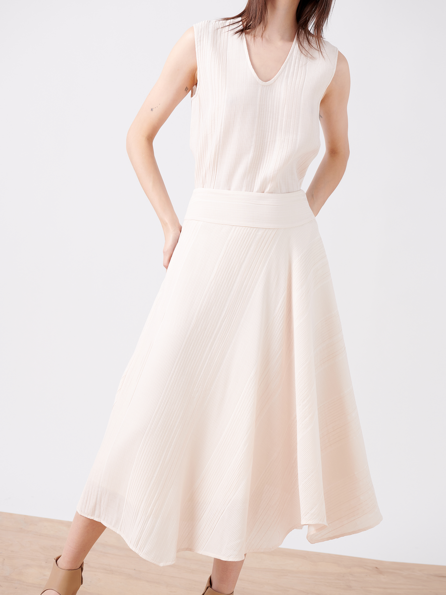 In a minimalist setting, a person models the Aki Wave Skirt - Final Sale by Zero + Maria Cornejo. Wearing a sleeveless, textured white top with a sleek sateen finish skirt that flows to mid-calf, they exude elegance while posing with one hand on their hip.